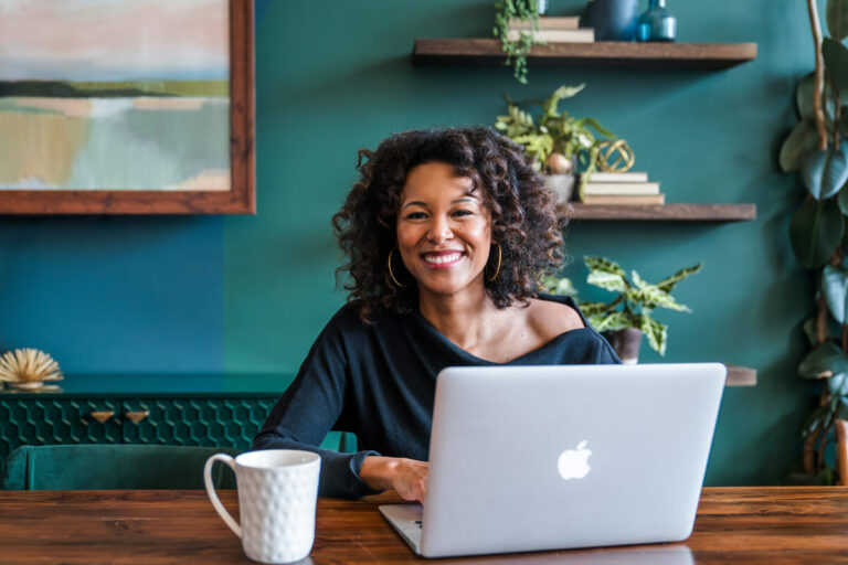 Woman with curly hair and gold hoop earrings smiling while using MacBook at wooden table, teal wall with shelves behind her, coffee mug nearby.
