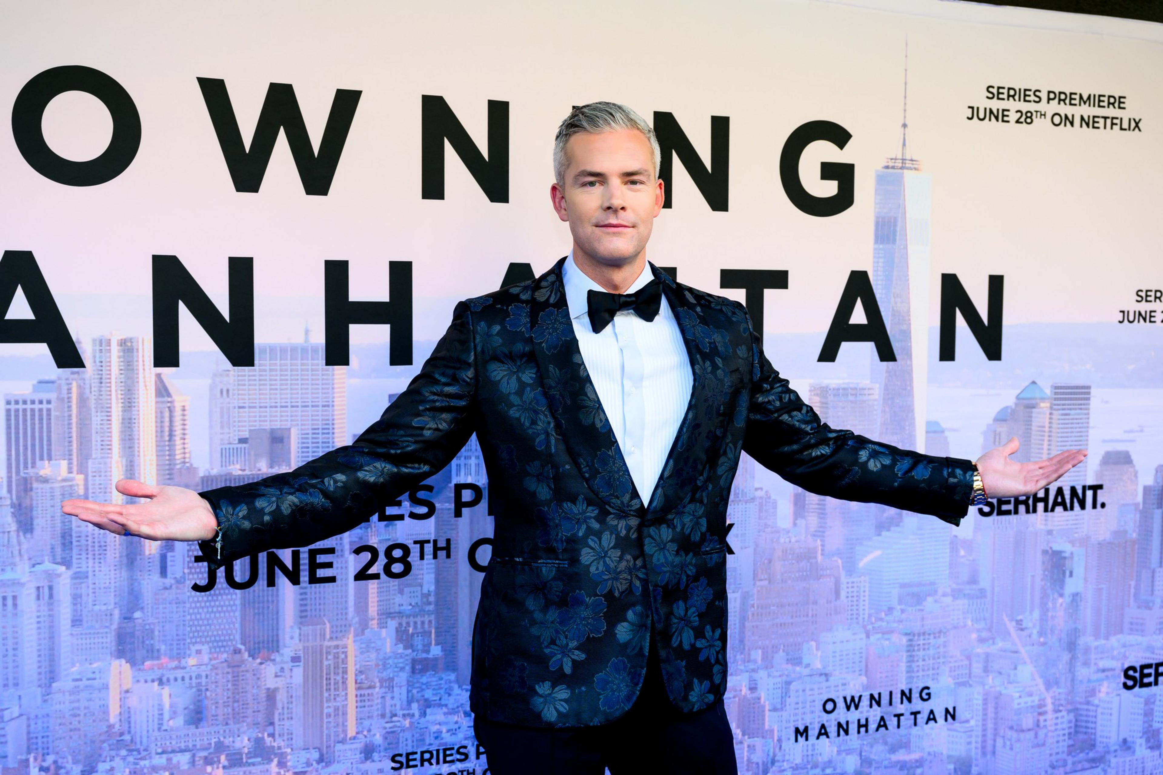 A man in a floral black tuxedo with a bow tie stands with arms outstretched in front of a backdrop featuring a city skyline and the words "OWNING MANHATTAN."