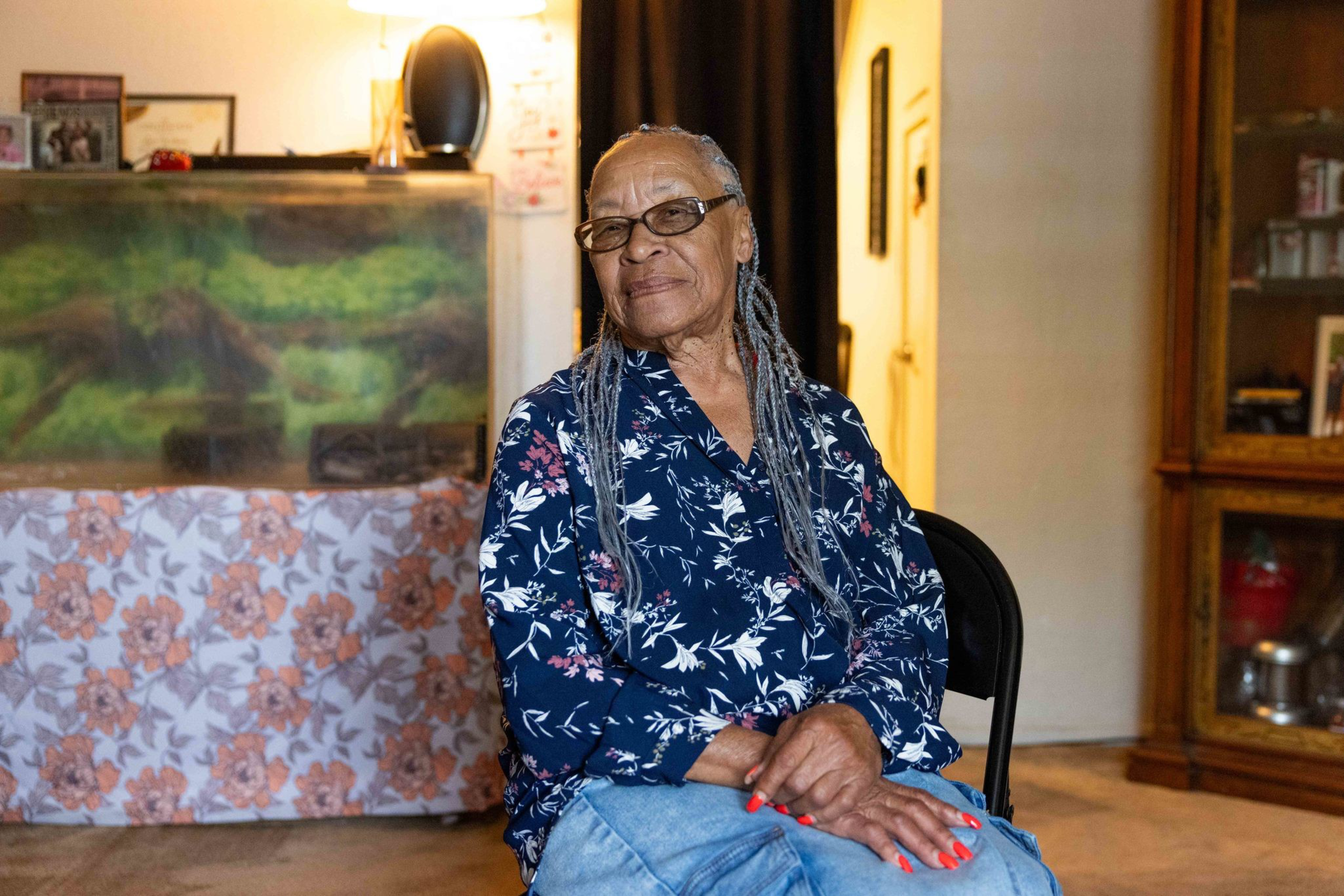 An older woman with long gray braids and glasses sits on a chair, wearing a blue floral shirt and light blue jeans, with a softly lit, warm room behind her.