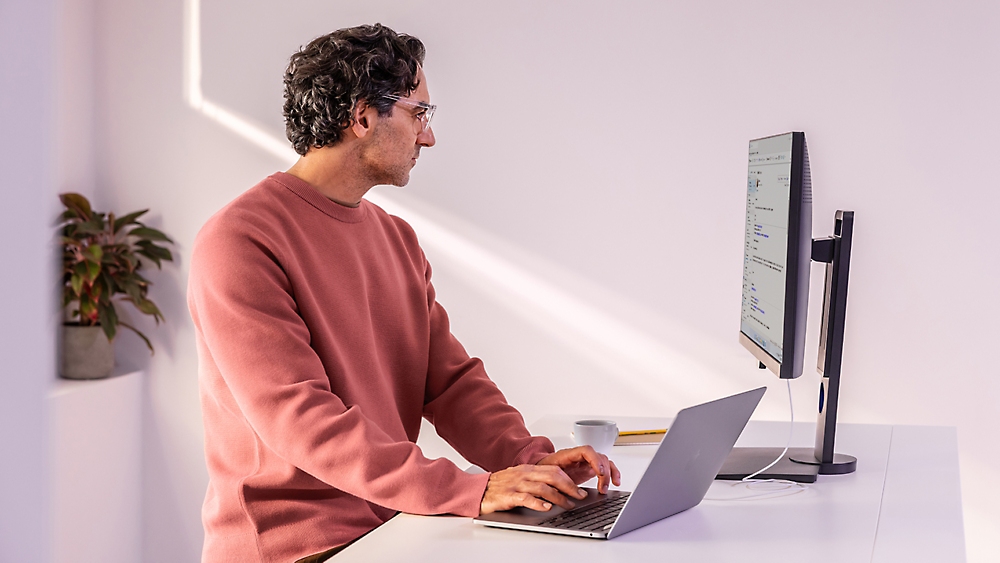 A man in a red sweater sitting at a desk typing on a laptop.