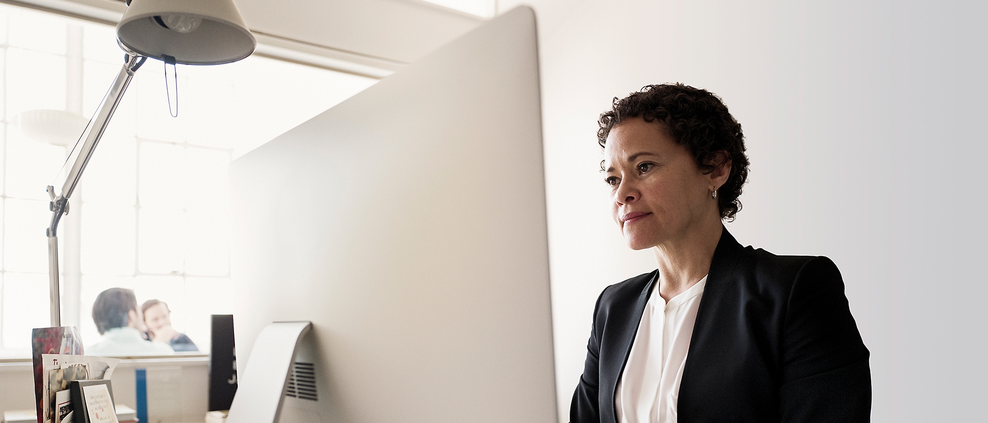 Nainen virallisissa vaatteissa työskentelee pöytätietokoneella valoisassa, modernissa toimistotilassa.