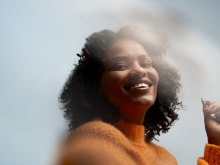 Une personne aux cheveux bouclés portant un pull-over orange sourit à la caméra.