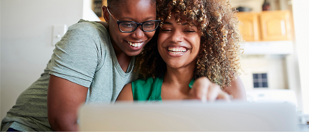 Two women laughing and embracing while looking at a laptop in a bright, homey room.