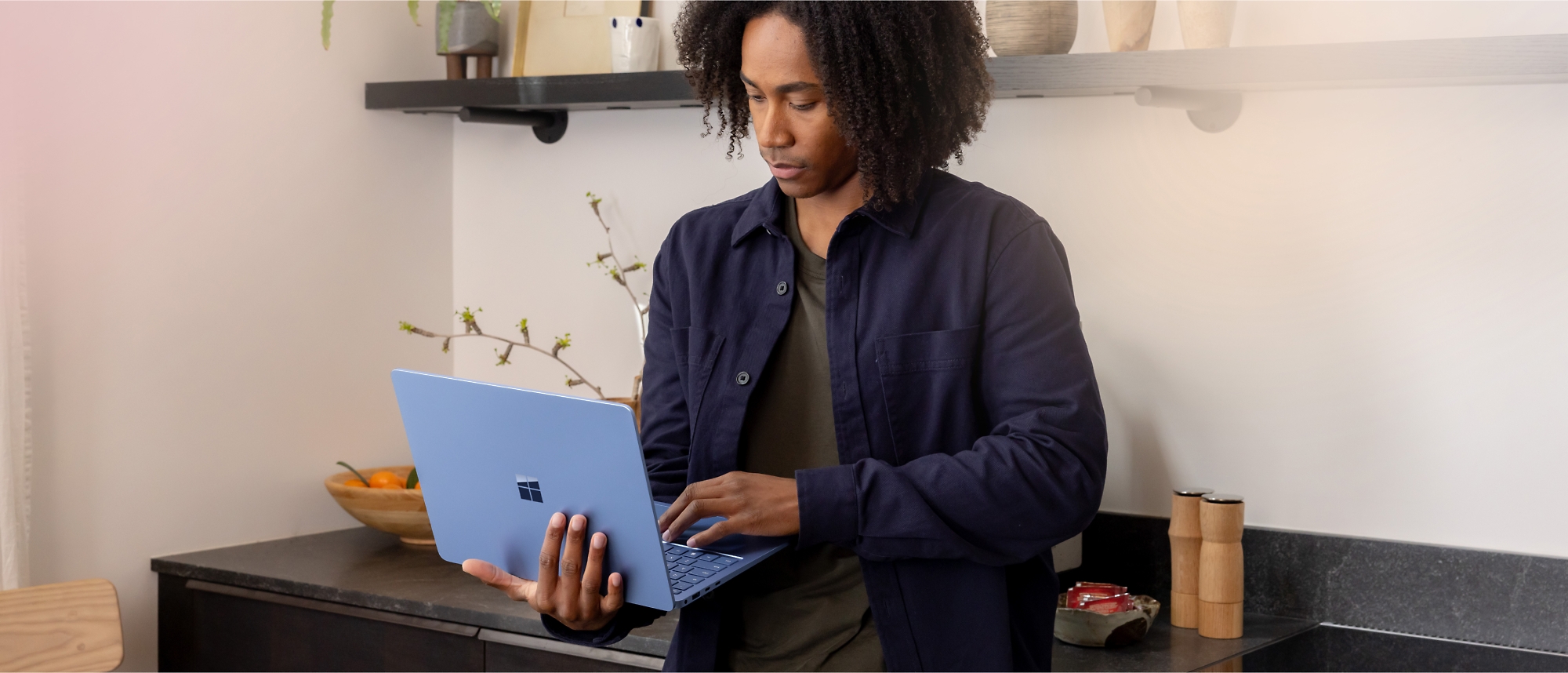 Person standing in a kitchen with a light blue laptop, focused on its screen. 