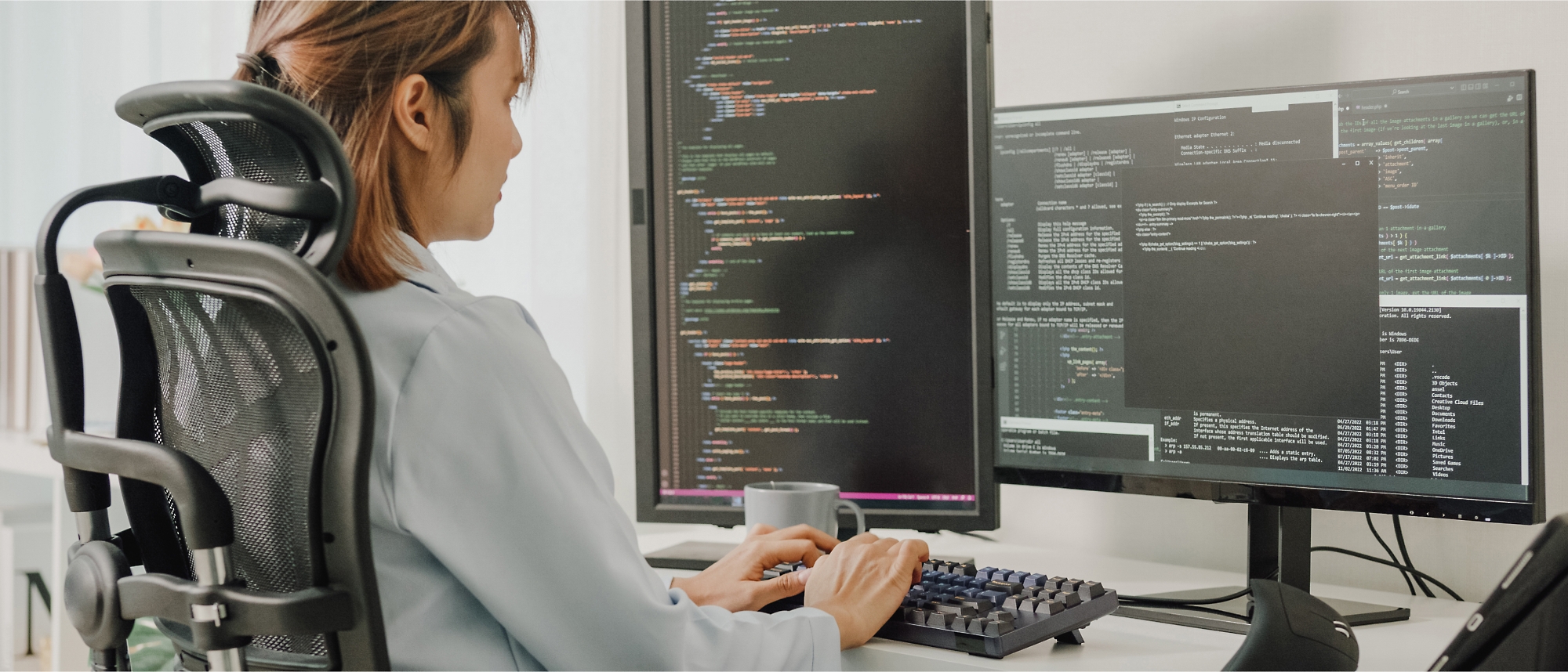 A person seated at a desk works on coding using two monitors displaying colorful programming text.