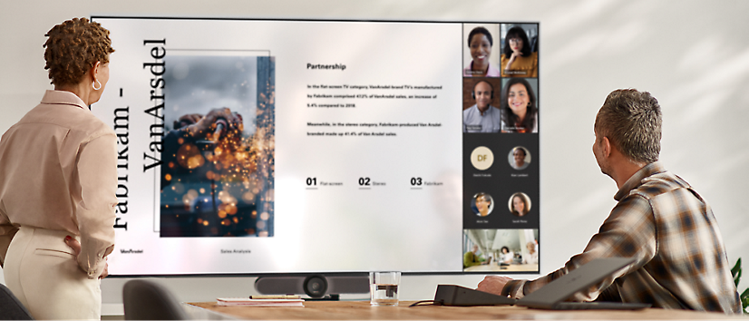 Two people are standing in a meeting room looking at a large screen displaying a virtual meeting 