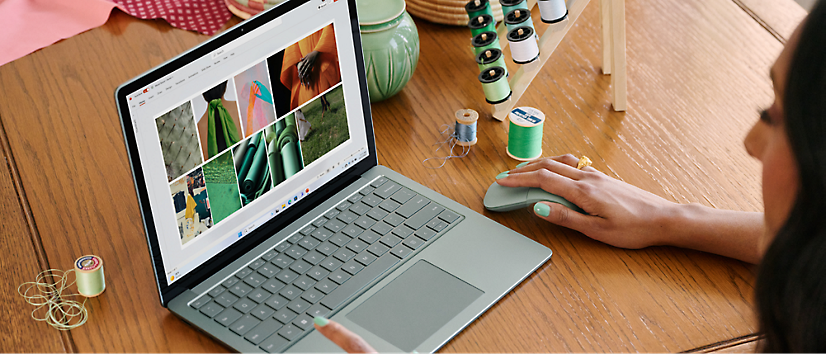 A person with green nails uses a laptop on a wooden table, surrounded by colorful spools of thread and sewing materials.