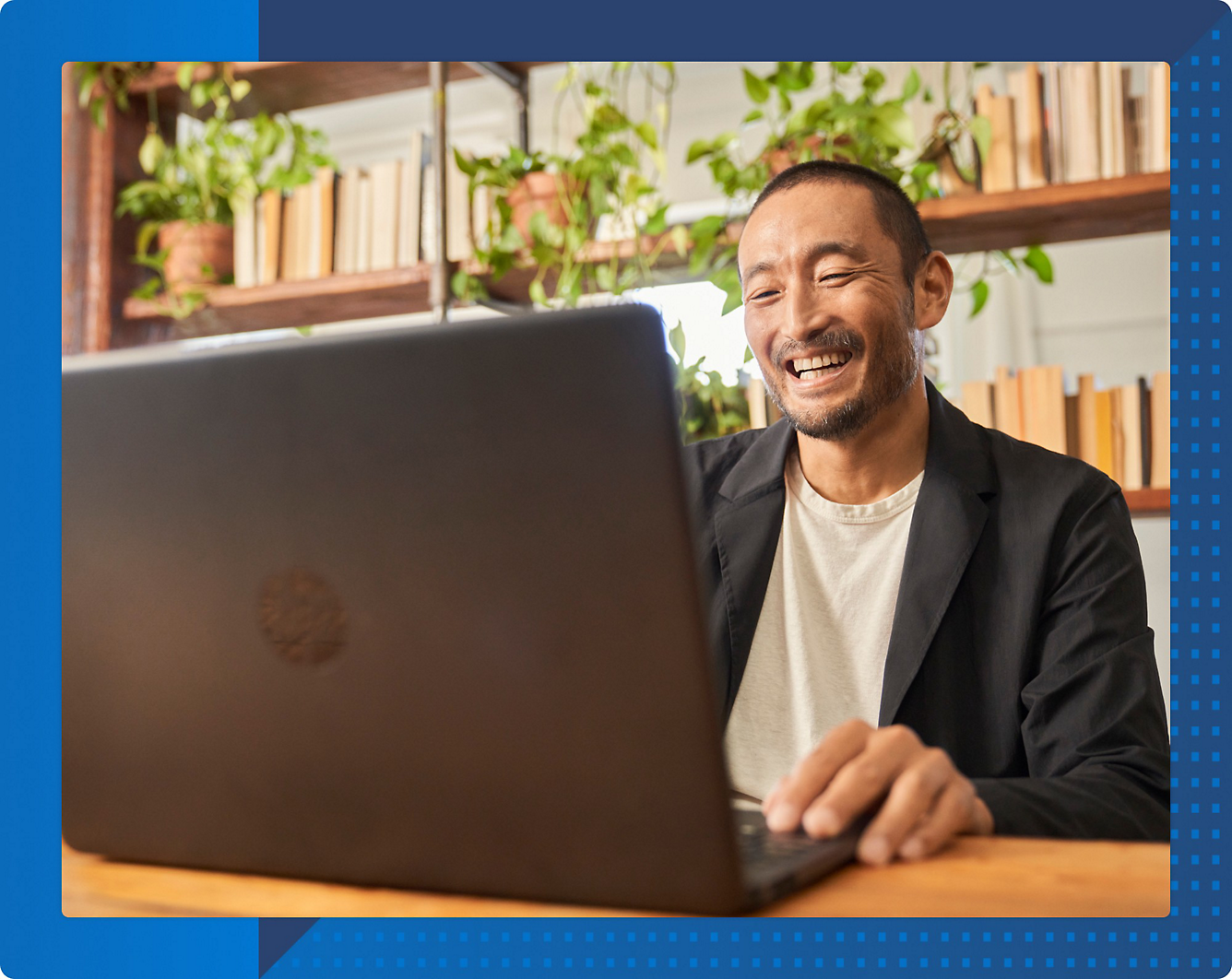 A person smiling while working on a laptop at a table, with shelves of books and potted plants in the background.