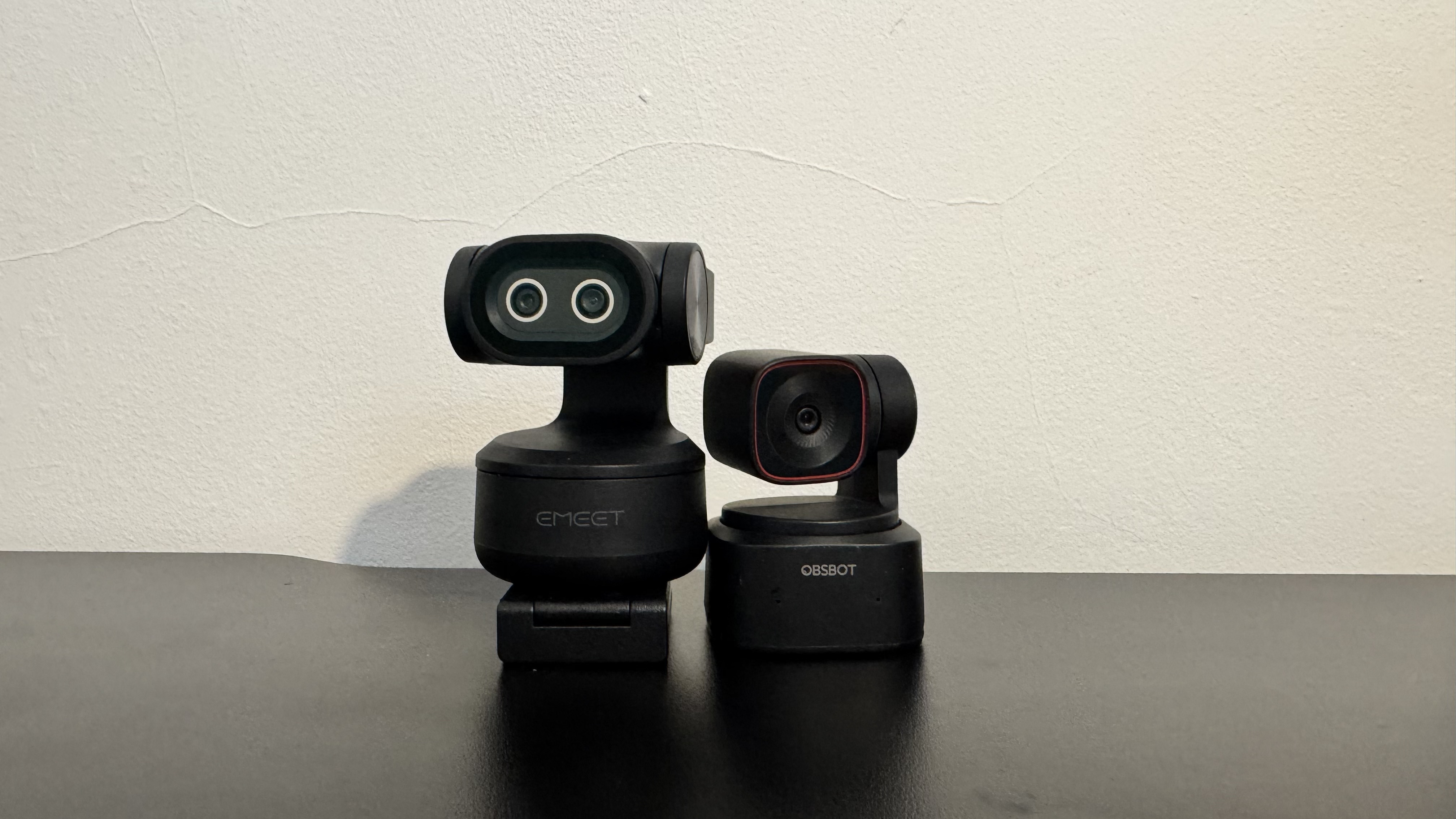 A side by side comparison of the Obsbot Tiny 2 Lite and the Emeet Pixy on a black desk. 