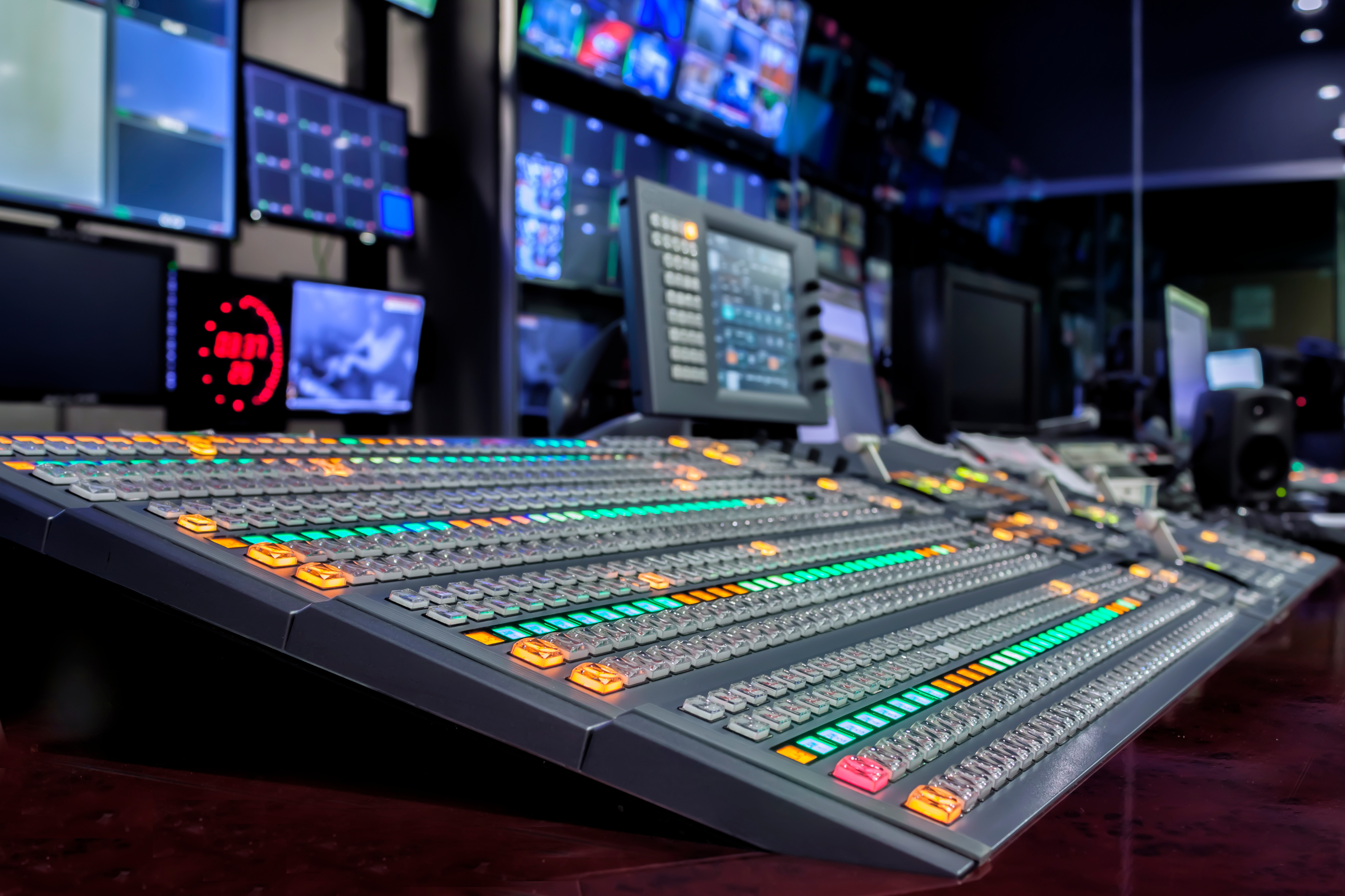 A close-up view of an advanced audio and video mixing console in a broadcast control room, with illuminated buttons and multiple display monitors in the background.