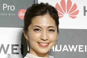 女性がレースのドレスを着てスマートフォンを持ち、カメラに向かって微笑んでいる。バックに企業ロゴが見える。