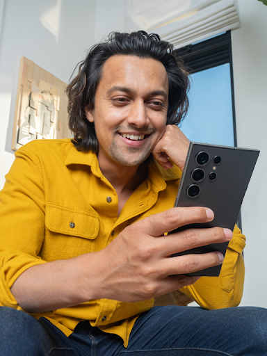 A man with dark, wavy hair and a yellow shirt smiles while using a smartphone. The background is blurred, suggesting a home or casual office setting.