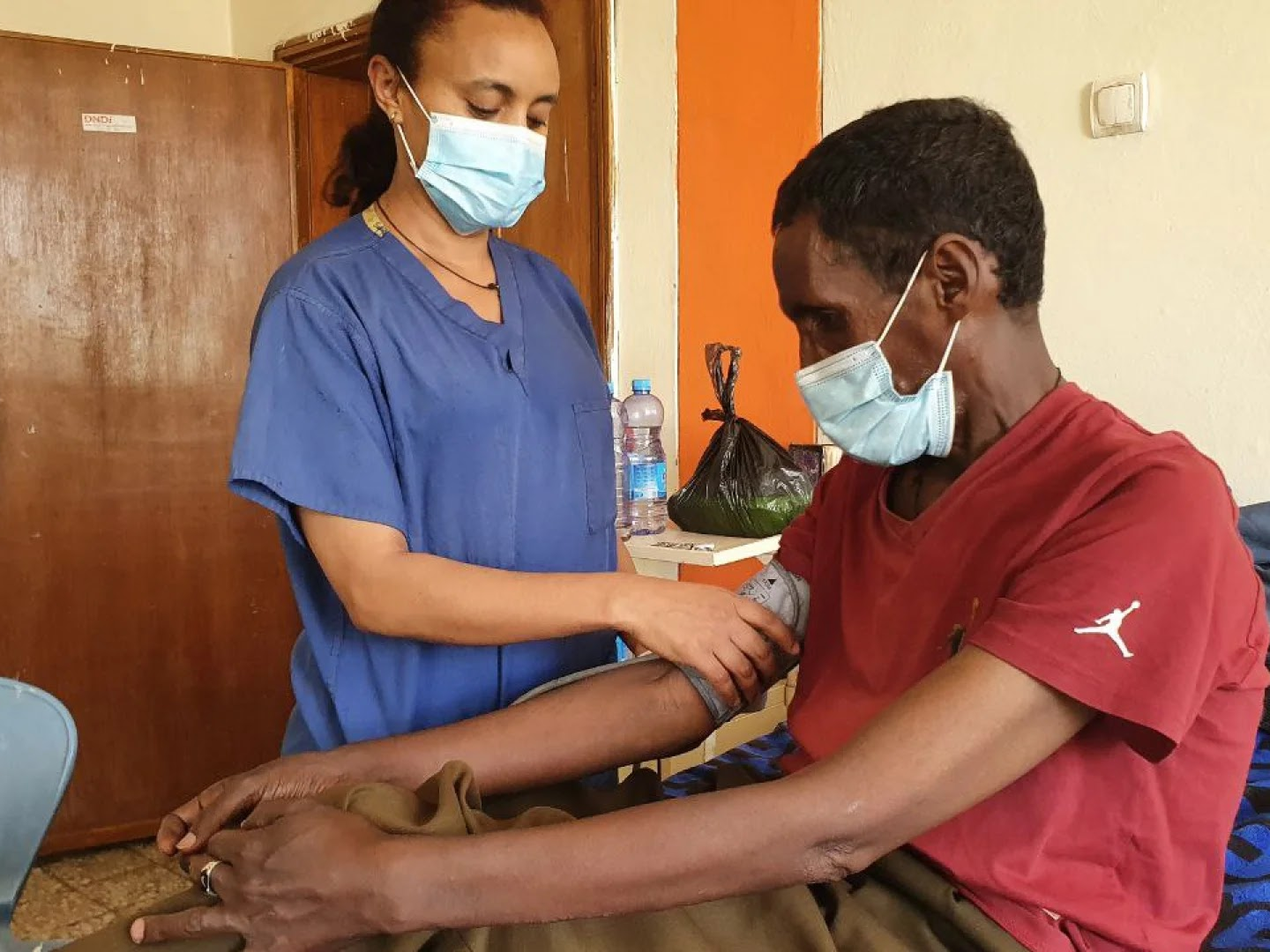 A healthcare worker in blue scrubs taking a patient's blood pressure in a clinical setting.