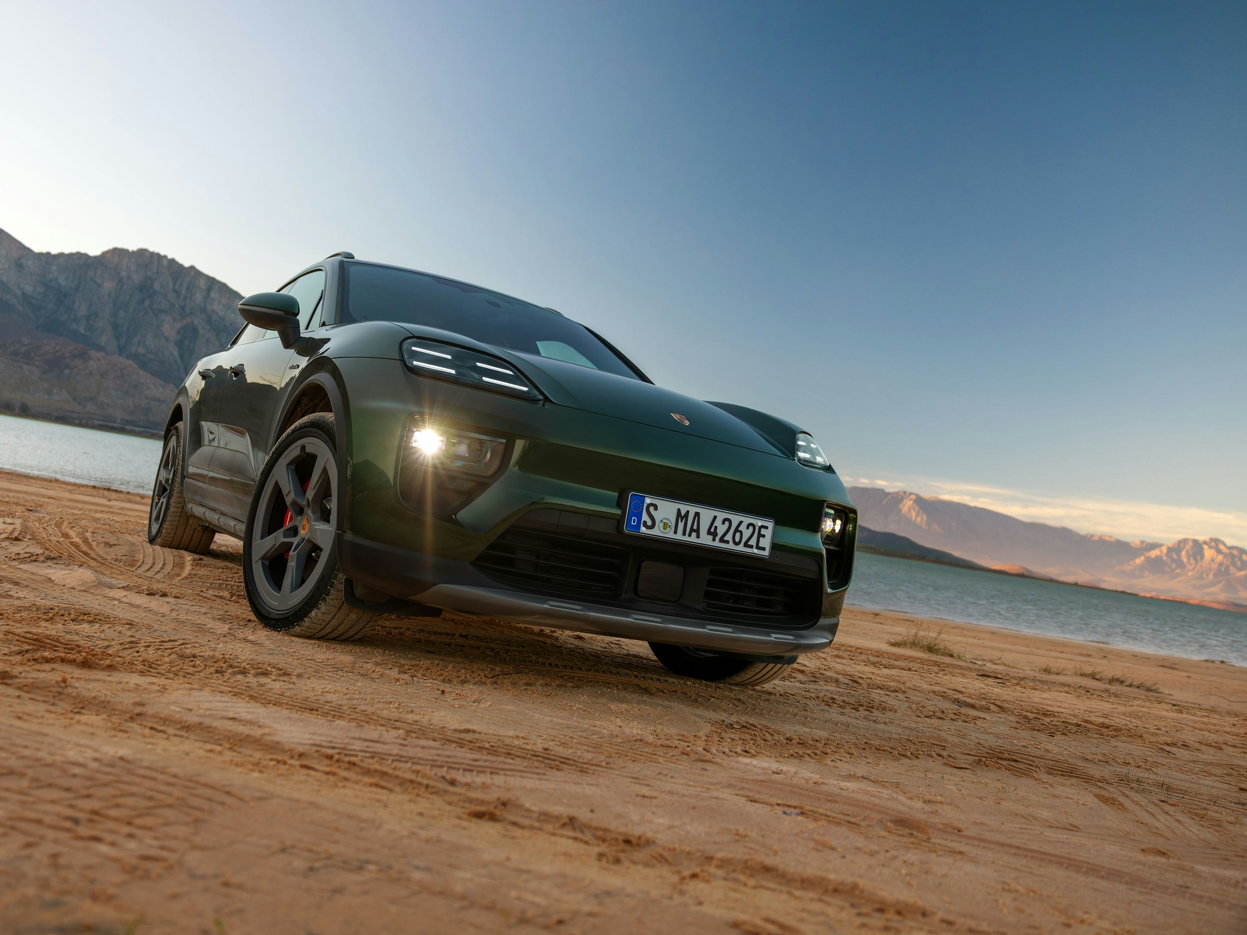 An Oak Green Metallic Neo Macan 4S standing in front of a lake with the mountains in the background.