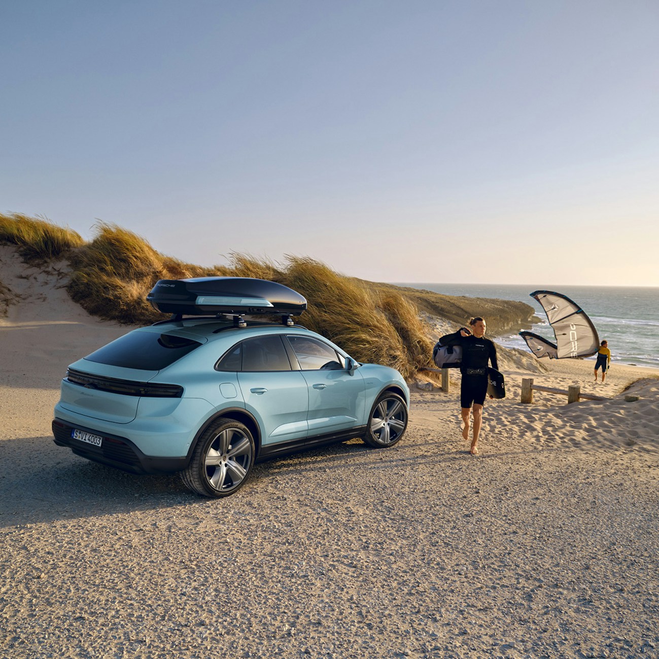 A man is walking on the beach next to a parked Porsche Macan 4 with a roof black box.
